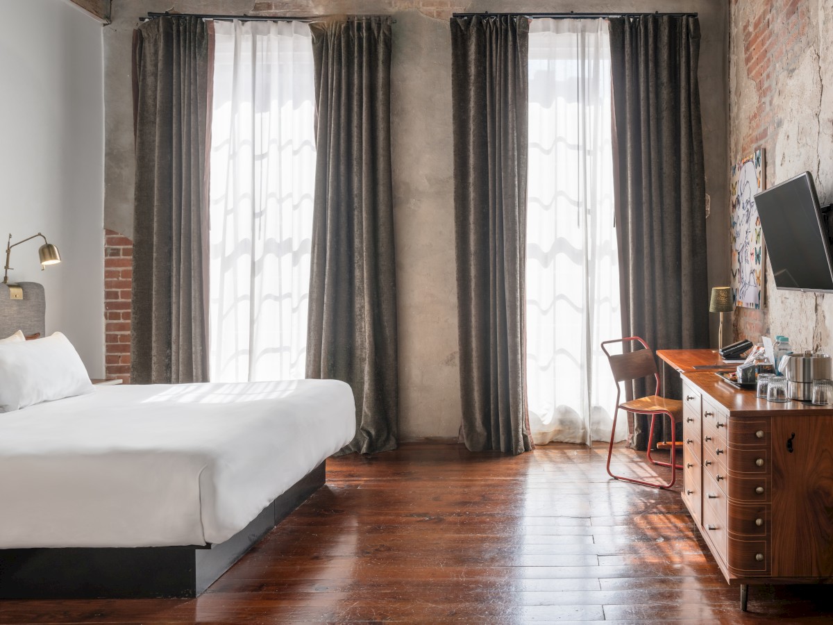 A stylish bedroom with a large bed, dark curtains, a flat-screen TV, wooden floors, and a desk with a chair by the window.