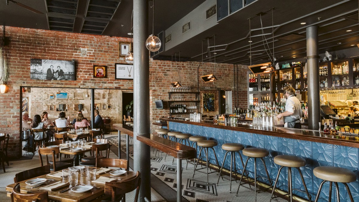 The image shows a cozy restaurant with a bar, stools, tables, and people dining, creating a warm and inviting atmosphere.