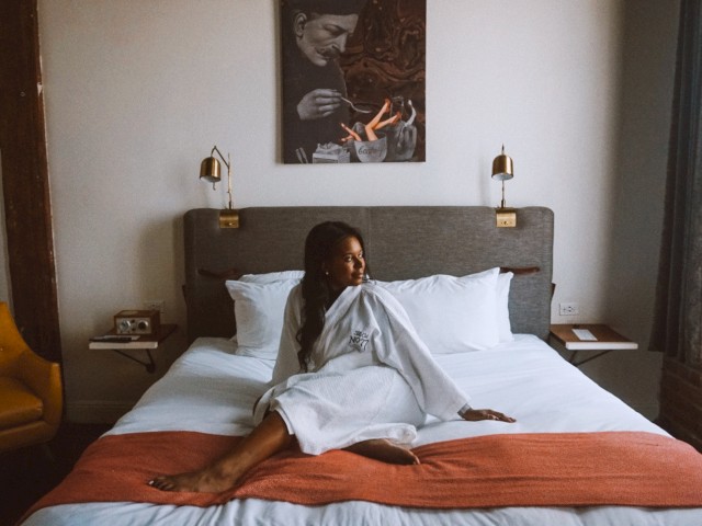 A woman wrapped in a towel sits on a bed with white linens and an orange throw, in a cozy room with modern decor and soft lighting.