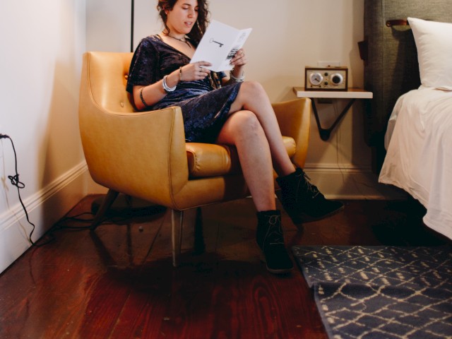 A person sitting in a yellow chair reading a book next to a bed in a well-lit room with minimalist decor, including a lamp and a small side table.