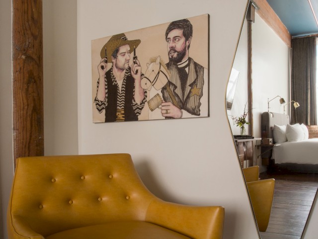 The image shows a room with a yellow chair, a mirror reflecting a bed, and a decorative painting of two figures on the wall.