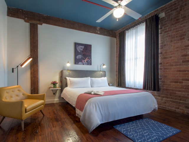 A cozy bedroom with a bed, yellow chair, artwork, floor lamp, and exposed brick and wood features. Ceiling fan and large window with curtains.
