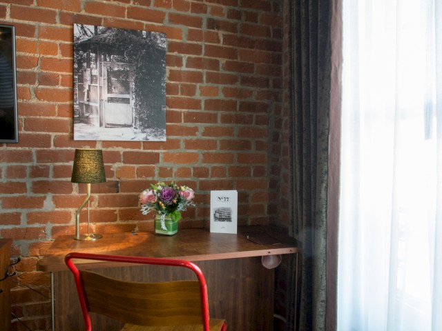 A cozy corner with a wooden desk, red chair, a lamp, a vase of flowers, a framed photo on the brick wall, and a window with sheer curtains.