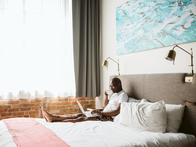 A person is sitting on a bed, using a laptop and drinking from a cup. The room has brick walls, curtains, and a colorful painting above the headboard.