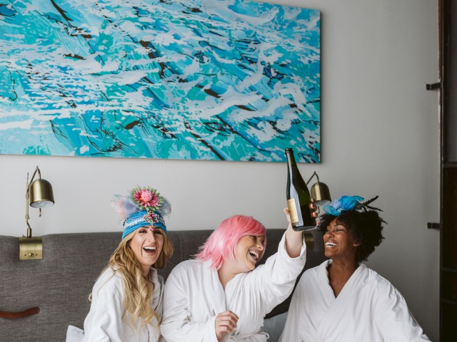 Three people wearing robes and feathered headbands are sitting on a bed; one holds up a bottle, and they are all smiling.