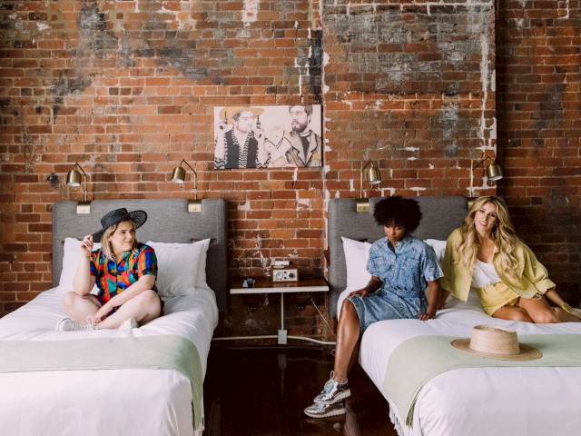 Three people sitting on two beds in a rustic room with exposed brick walls and a photo on the wall above them, ending the sentence.