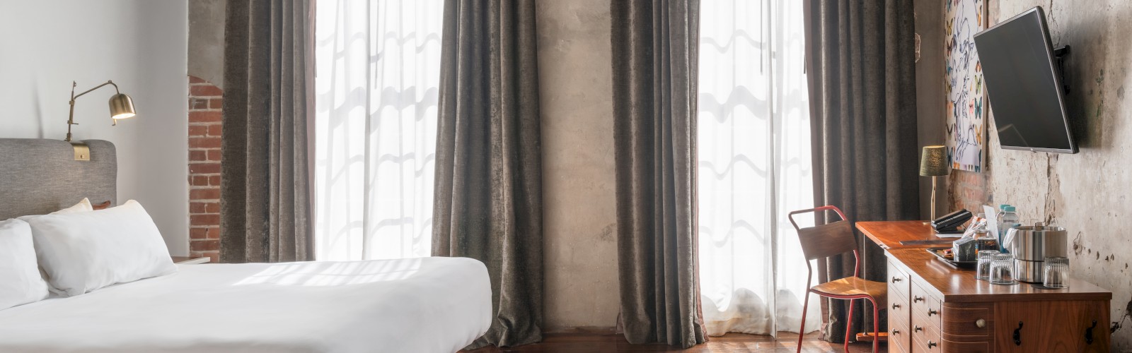 A modern hotel room features a large bed, a wooden desk with a chair, a wall-mounted TV, and two tall windows with dark curtains.