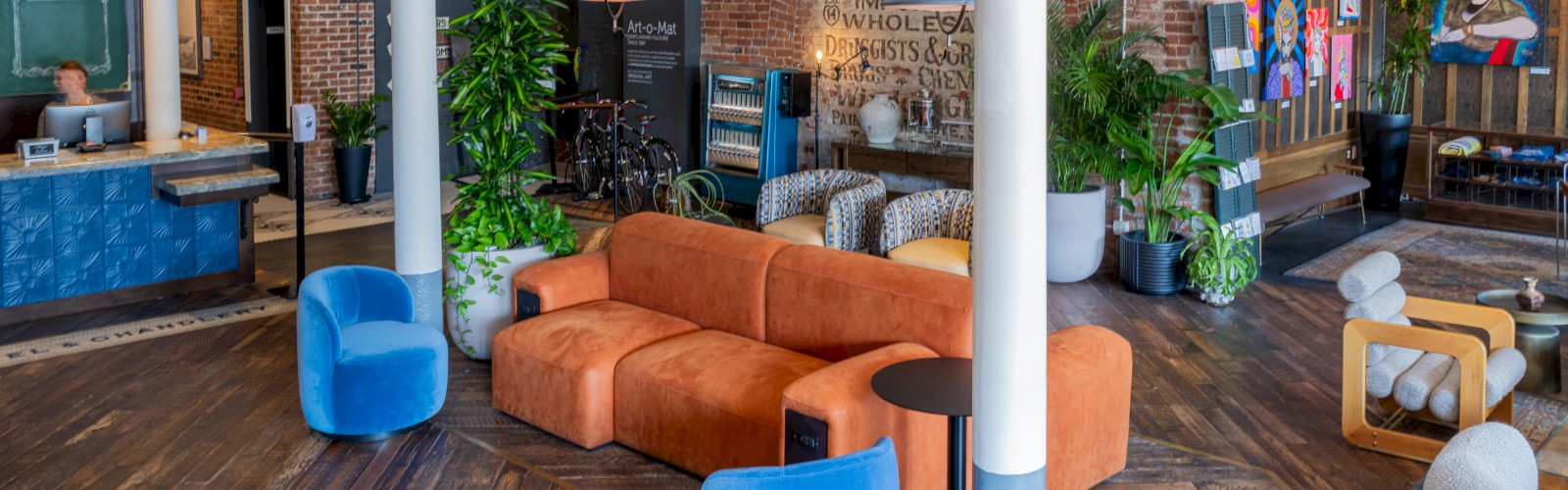 A modern lounge area with orange and blue seating, plants, a reception desk, and artwork on brick walls, featuring stylish lighting and wooden flooring.