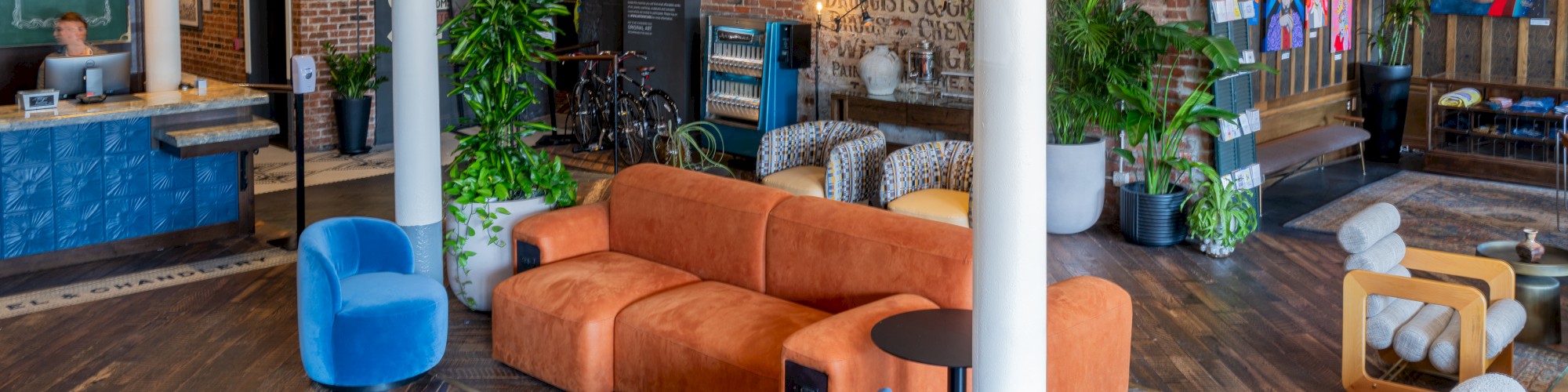 A modern lounge area with orange and blue seating, plants, a reception desk, and artwork on brick walls, featuring stylish lighting and wooden flooring.