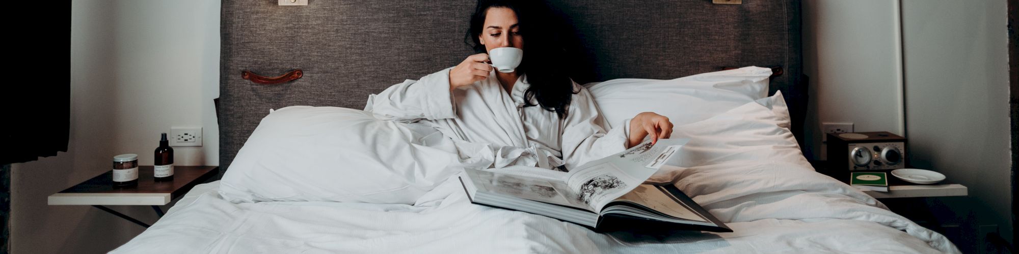 A person sits in bed drinking from a cup and reading a magazine, under modern art, with bedside lights turned on.