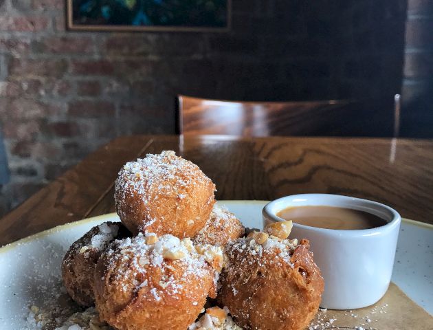 The image shows a plate with several powdered sugar-covered desserts, possibly fritters or doughnuts, garnished with nuts and a small cup of sauce.