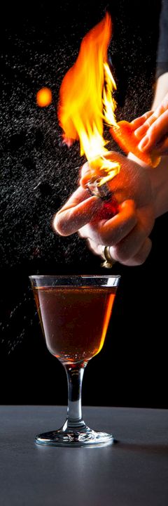 A person is igniting something and creating a burst of flame above a drink in a cocktail glass, likely performing a flaming cocktail trick.