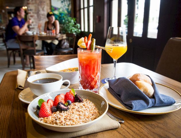 The image shows a breakfast setting with granola and fruit, a cup of coffee, a Bloody Mary, an orange juice, and bread rolls in a blue napkin.