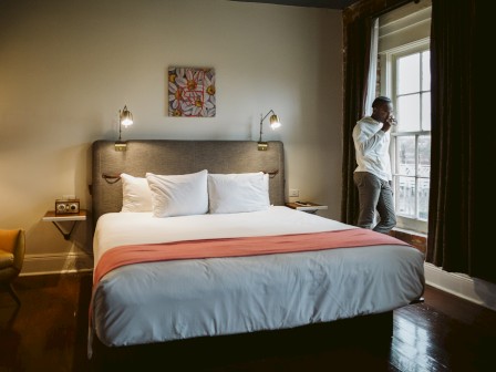 A cozy bedroom with a large bed, white and pink bedding, a man standing by the window, a small wall painting, bedside lamps, and a chair.