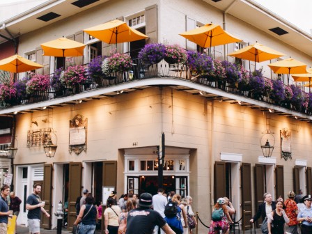 People walk and cycle past a corner building with an outdoor balcony adorned with flowers and orange umbrellas.