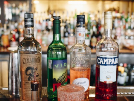 The image shows a bar setup featuring bottles of various liquors, a cocktail in the foreground, and a background of additional bottles on shelves.