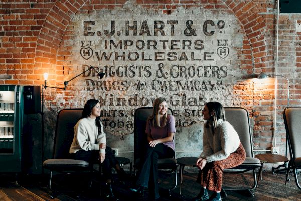 Three people are sitting and conversing in front of an old brick wall with a vintage advertisement for E.J. Hart & Co. Importers & Wholesale.