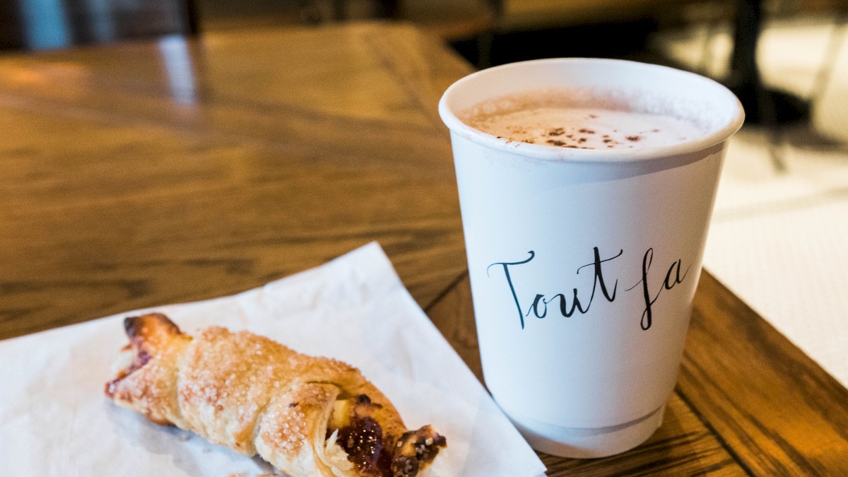 A croissant pastry on a white paper and a cup of coffee labeled 