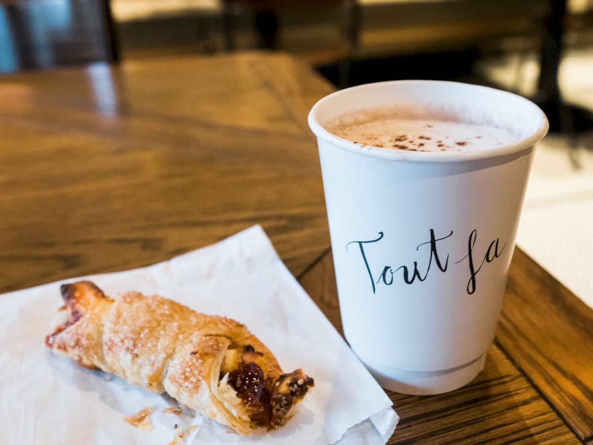 A croissant pastry on a white paper and a cup of coffee labeled 