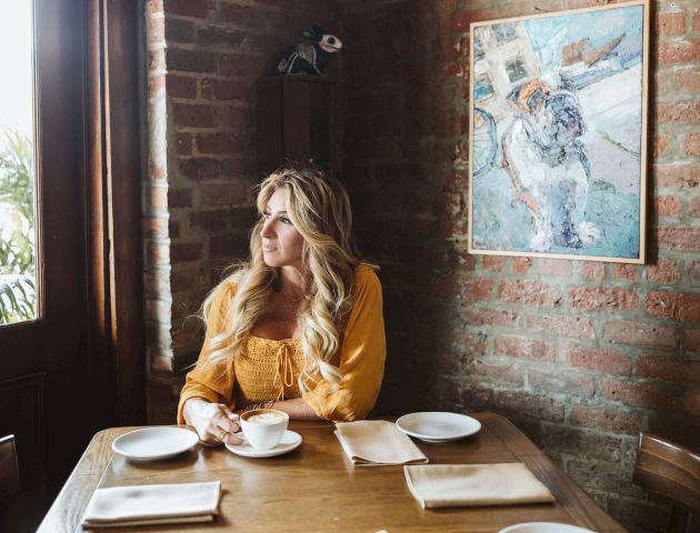 A woman in a yellow top sits at a wooden table in a brick-walled cafe, holding a cup of coffee and looking out the window, with a painting on the wall.