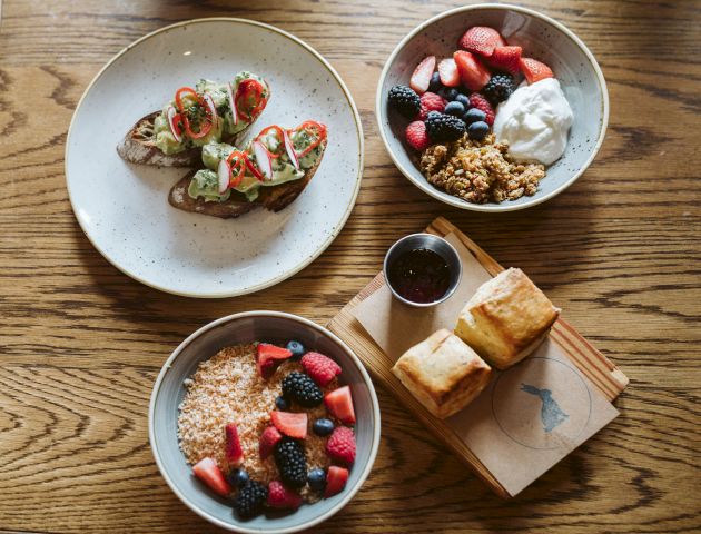 The image shows a breakfast spread with avocado toast, yogurt with granola and berries, and a bowl of mixed berries, alongside two pieces of bread and jam.