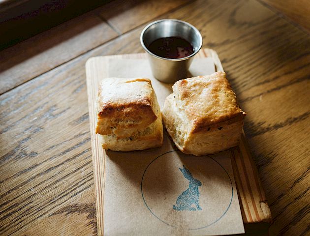 The image shows two square pieces of bread or pastry on a wooden board near a window, accompanied by a small metal cup with a dark dipping sauce.