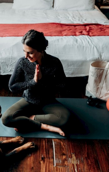 A woman sits on a yoga mat in a bedroom, hands in prayer position, facing a dog. A tote bag and a red ball are nearby, ending the sentence.