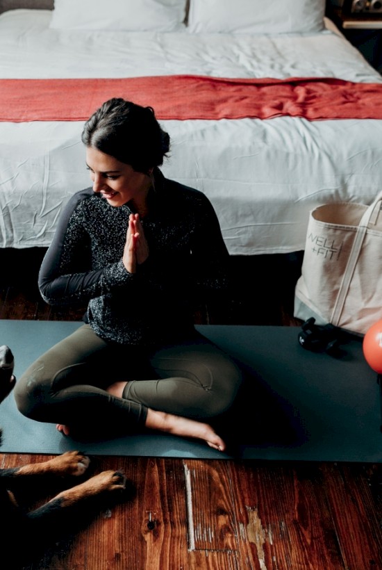 A woman sits on a yoga mat in a bedroom, hands in prayer position, facing a dog. A tote bag and a red ball are nearby, ending the sentence.