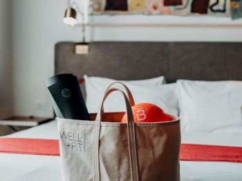 A tote bag containing workout gear, including a yoga mat and an orange item, is placed on a table in front of a neatly made bed in a stylish room.