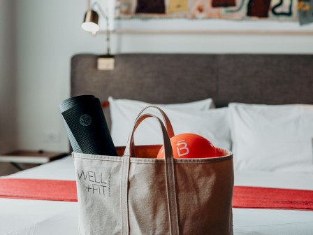 A tote bag containing workout gear, including a yoga mat and an orange item, is placed on a table in front of a neatly made bed in a stylish room.