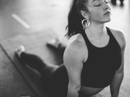 A woman in a gym is performing the upward dog yoga pose in black and white. Fitness equipment is visible in the background.