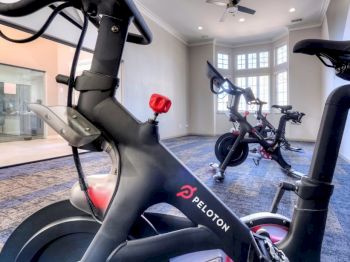 The image shows a room with two Peloton exercise bikes on a carpeted floor. There is a ceiling fan and large windows in the background.