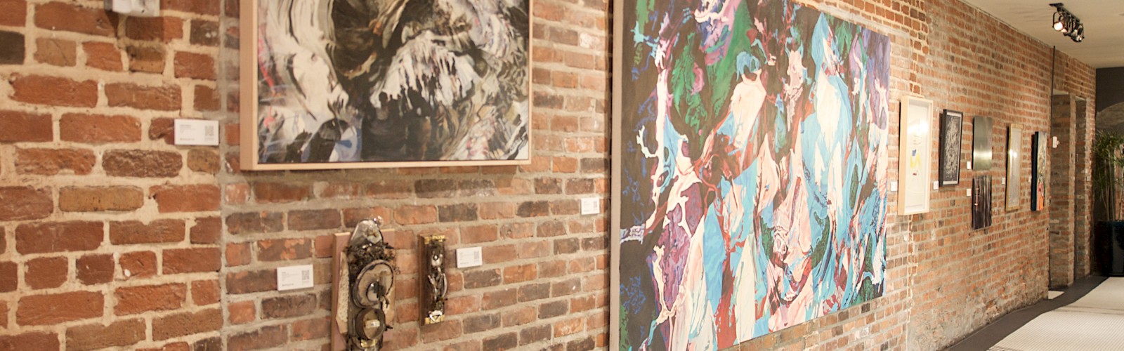 The image shows an art gallery with various paintings hanging on a brick wall and a well-lit hallway.
