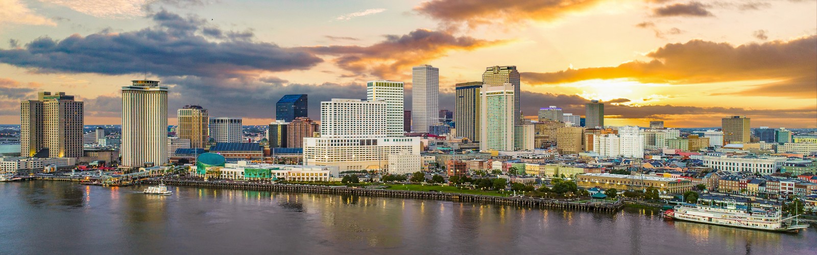 A coastal city skyline at sunset with tall buildings along a waterfront, reflecting warm orange-pink skies over calm water.
