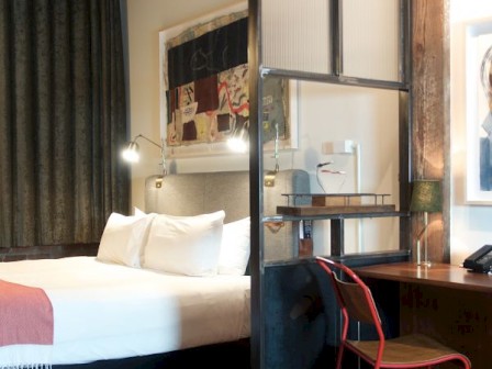 A cozy bedroom with a bed, desk, chair, and a partition. The room features modern decor with artwork, a lamp, and curtains in the background.