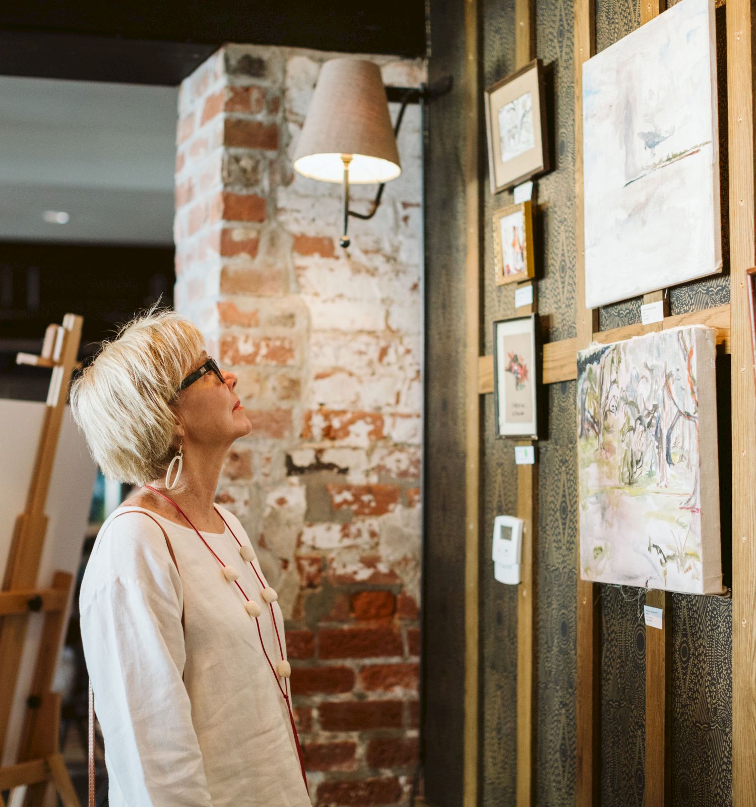 A person with short blonde hair and glasses is looking at framed artworks on a wall in a rustic-themed gallery or café.