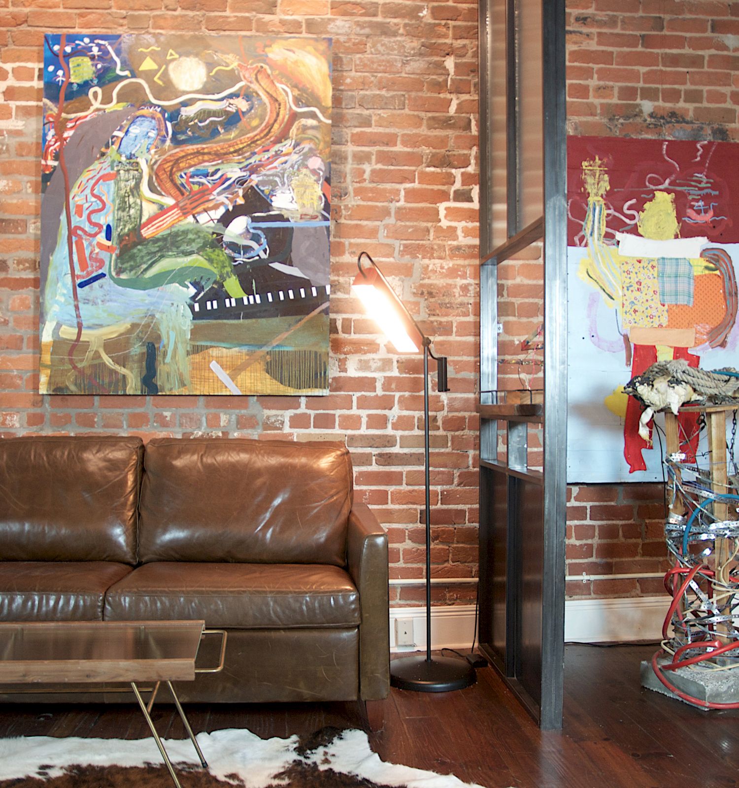 A modern living room with exposed brick walls, abstract art, a leather sofa, a sleek coffee table on a cowhide rug, and a unique floor lamp.