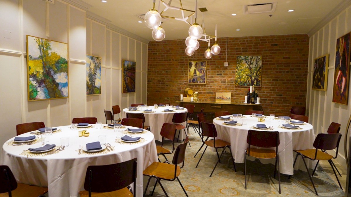 The image shows an elegant dining room with round tables, white tablecloths, blue napkins, paintings on the walls, and modern lighting.