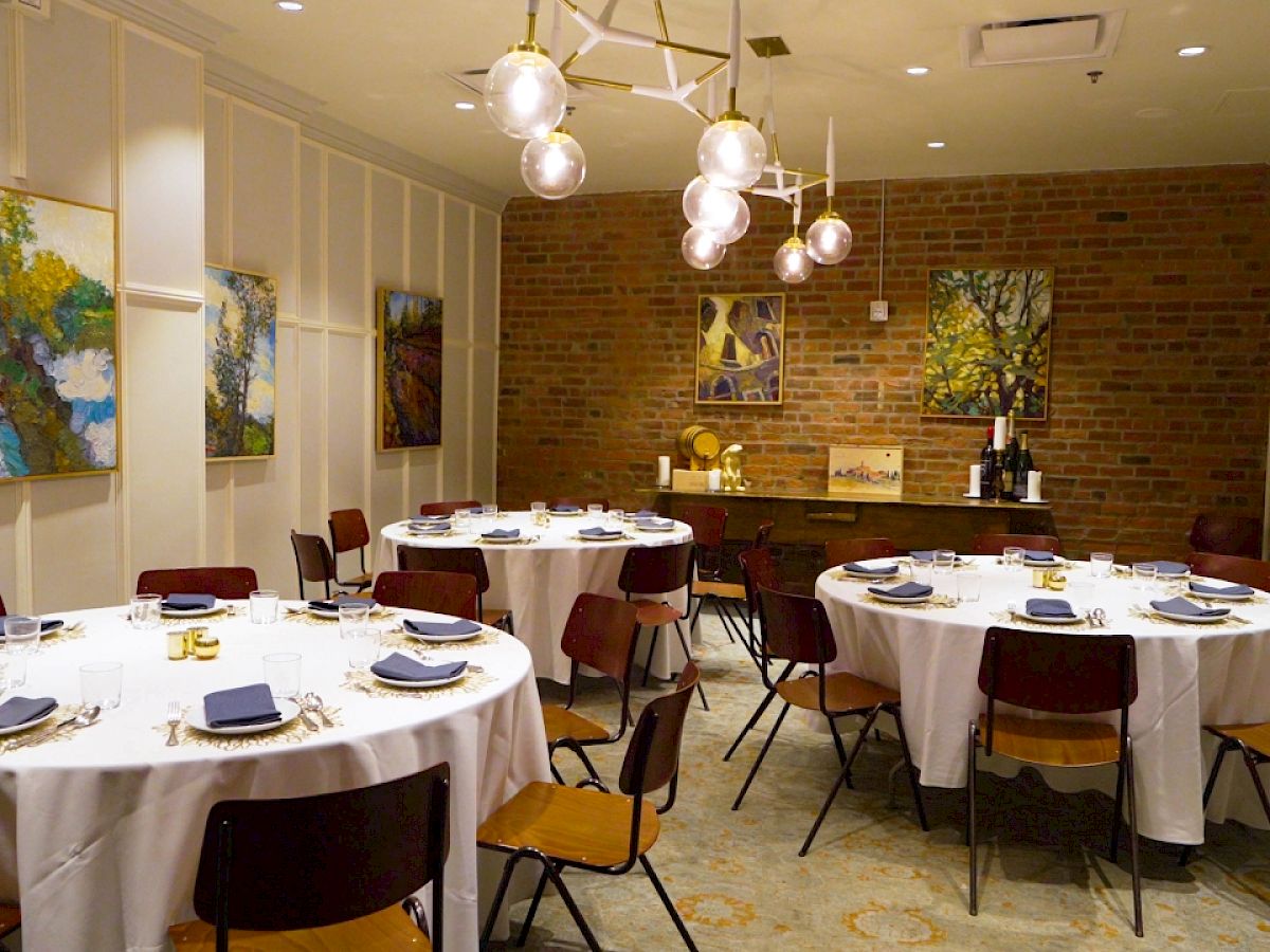 The image shows an elegant dining room with round tables, white tablecloths, blue napkins, paintings on the walls, and modern lighting.