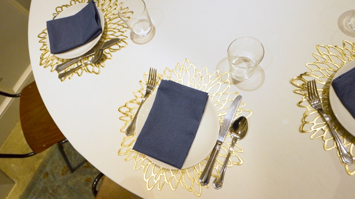 The image shows a table set for a meal, with plates, navy blue napkins, cutlery, and glasses on decorative gold placemats in a floral pattern.