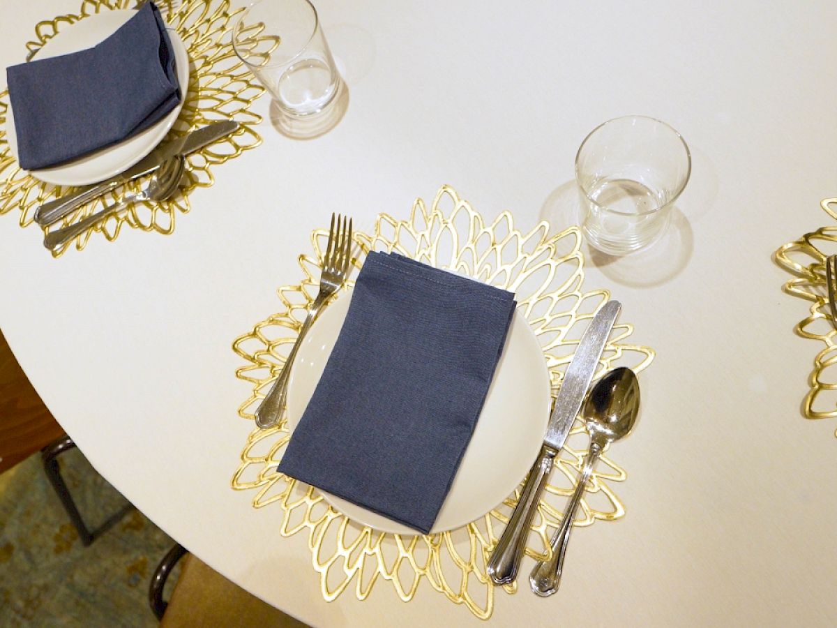 The image shows a table set for a meal, with plates, navy blue napkins, cutlery, and glasses on decorative gold placemats in a floral pattern.