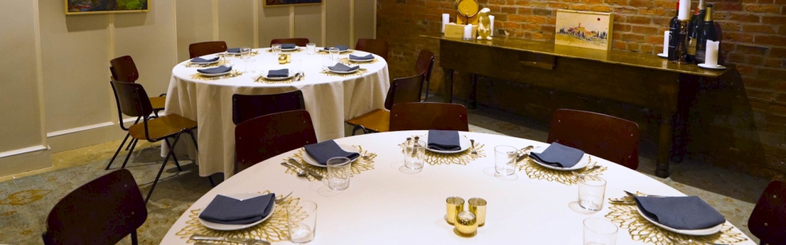The image shows a dining room with round tables set with plates and napkins, four paintings on the walls, and a sideboard with bottles and decor items.