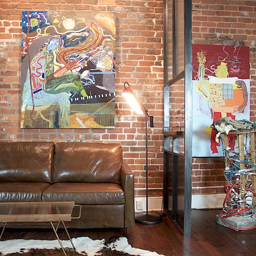 A living room features a brown leather sofa, abstract wall art, a modern coffee table, and an artistic sculpture against an exposed brick wall.