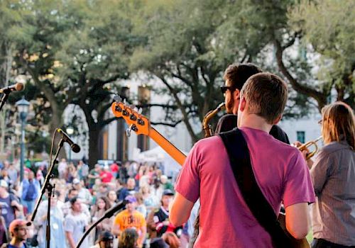 Band performing live on stage viewed from behind, facing an audience outdoors.