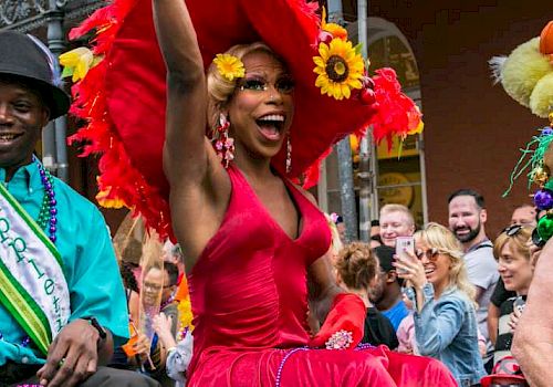 Three people in festive attire are on a gay LBGTI parade float, cheering with a crowd around