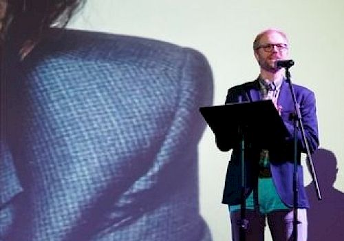 A man is presenting with a microphone; a large screen is in the background.