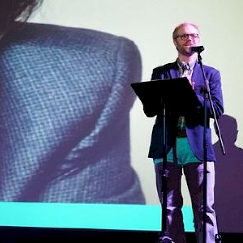 A man is presenting with a microphone; a large screen is in the background.