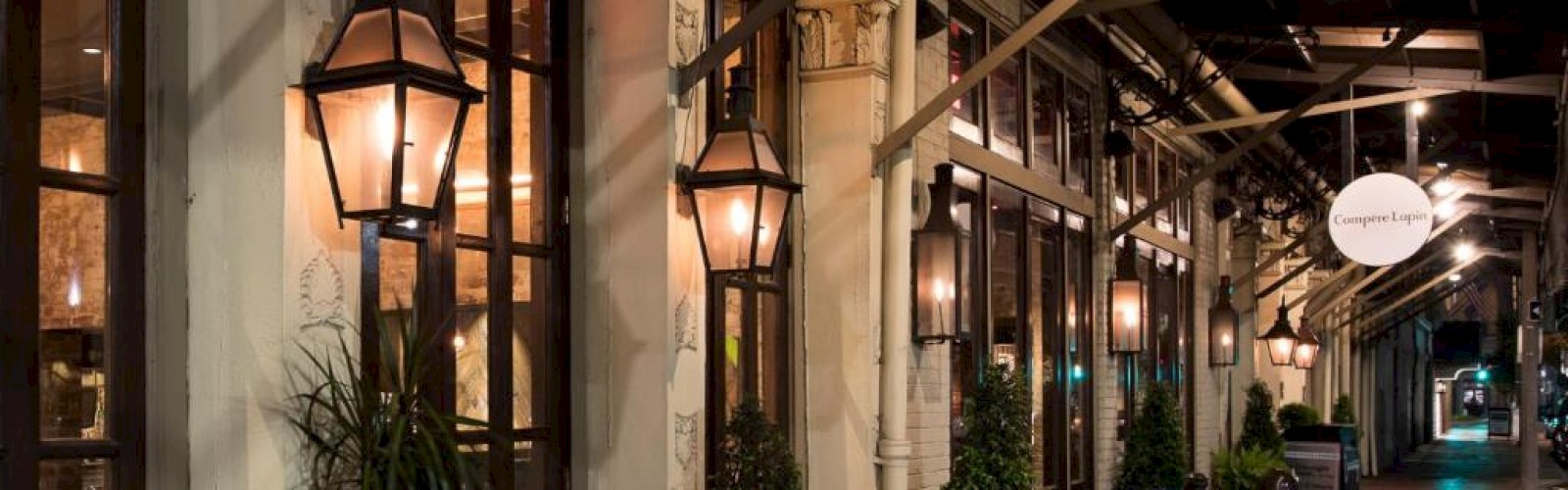 A cozy, illuminated street scene featuring multiple lanterns, potted plants, and a restaurant or shop with large windows and an awning.