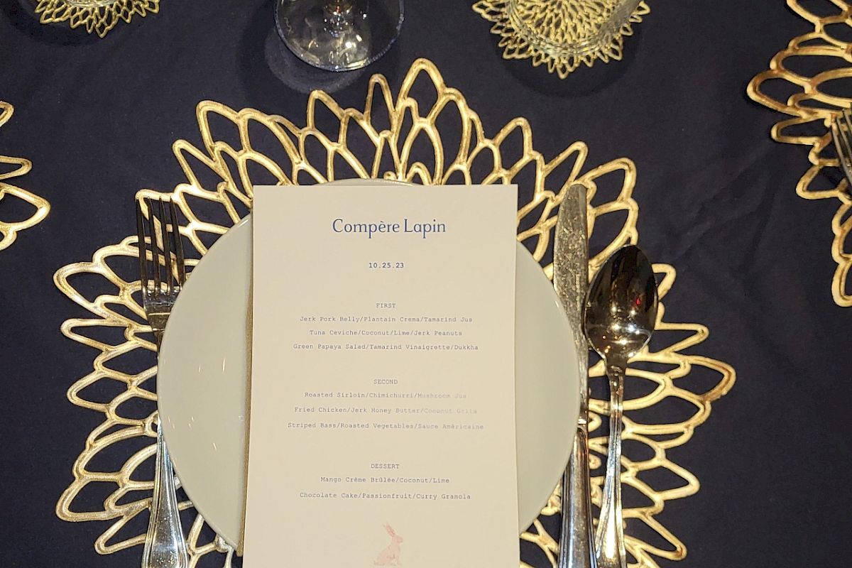 A table setting with a decorative placemat, plate, utensils, and glasses, featuring a menu on the plate atop a dark tablecloth, ending the sentence.