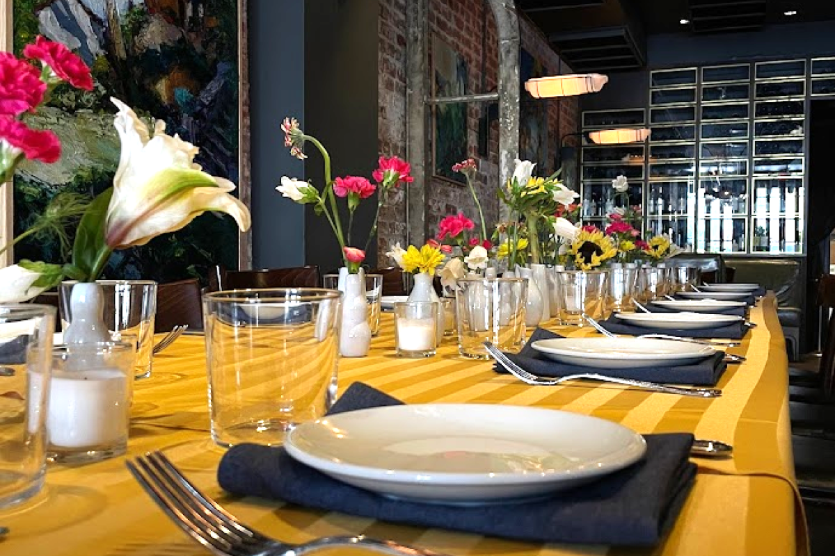 A long dining table with a yellow tablecloth, decorated with flowers, plates, glasses, and cutlery. The setting is in a stylish, modern restaurant.